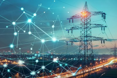 Auf dem Bild ist eine symbolische Darstellung eines intelligenten Stromnetzes (Smart Grid) zu sehen. Auf der rechten Seite steht ein großer Hochspannungsmast (Strommast) bei Dämmerung. Im Hintergrund erstreckt sich eine beleuchtete Stadtlandschaft mit Straßen und Gebäuden. Über das gesamte Bild – besonders auf der linken Seite – ist ein abstraktes, leuchtendes Netzwerk aus Linien und Datenpunkten (Knotenpunkten) gelegt.